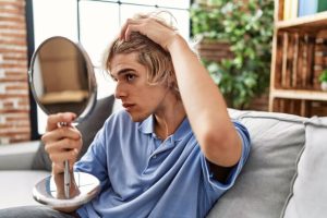 Jeune homme blond se regardant les cheveux dans le miroir, assis sur le canapé à la maison.