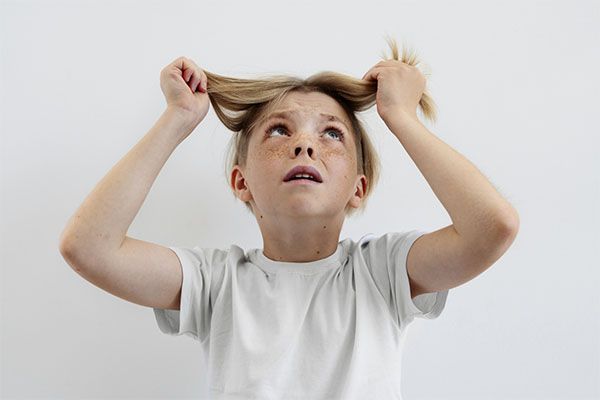 Un garçon blond en chemise blanche, l'air stressé, se prend les cheveux dans les mains. l'alopécie infantile