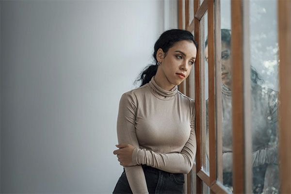 Femme en chemise grise au visage inquiet près de la fenêtre d'une maison. 
