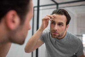 Apparence. Un jeune homme vêtu d'un tee-shirt gris se regarde dans le miroir- greffe de cheveux en France