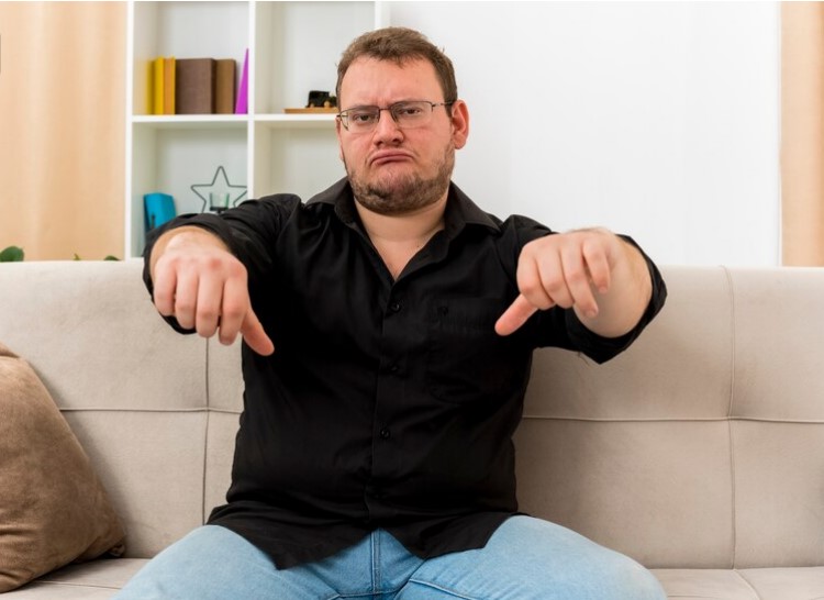 Homme adulte slave déçu avec des lunettes optiques, assis sur un fauteuil, pouces vers le bas avec les deux mains dans le salon.