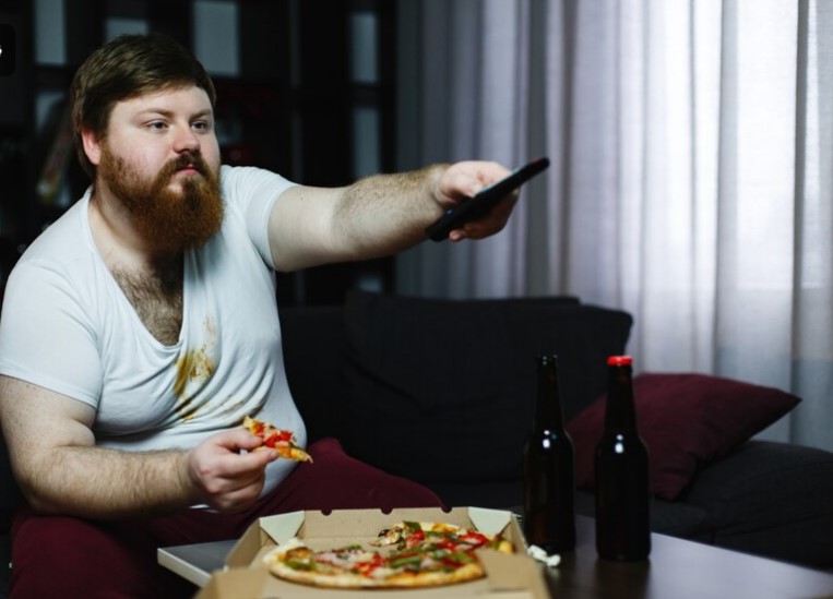 Le gros mange une pizza assis sur le canapé et change de chaîne sur le téléviseur.