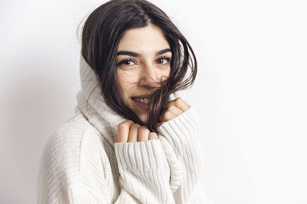 Belle femme en pull blanc, les mains près du visage et souriante. prendre soin de ses cheveux en hiver