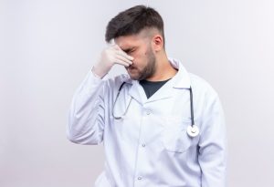 Un beau jeune médecin en blouse blanche, gants blancs et stéthoscope se tenant le nez qui coule, debout sur le mur blanc.