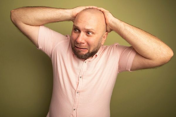 Concerned looking at side middle-aged bald man wearing pink t-shirt holding hands on behind head 