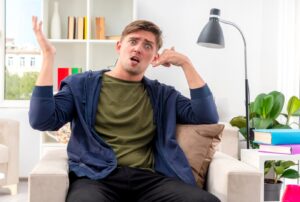 Un jeune homme blond, beau et enthousiaste, est assis dans un fauteuil et agite son téléphone en levant la main à l'intérieur du salon. vitamines pour prévenir l'alopécie