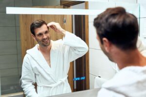 Reflet dans le miroir d'un homme heureux en peignoir qui ajuste sa coiffure.