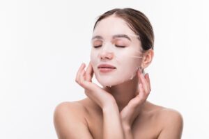 Soins du visage et traitements de beauté. Jeune femme avec un masque hydratant en tissu sur le visage, isolée sur un mur gris.