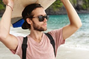 Hobby et vacances. Beau jeune homme barbu avec des lunettes de soleil stylées et une snapback, tenant une planche de surf au-dessus de sa tête, regardant vers l'océan, attendant les grosses vagues.