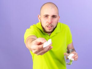 Un jeune homme caucasien pâle et malade tient un paquet de pilules médicales et un verre d'eau sur un fond violet.
